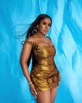 A woman in a golden Jacquard dress with intricate detailing, flower buds on the straps and hem, and a cinched waist. The dress shimmers with a metallic glow.